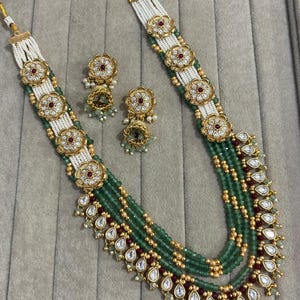 May include: An ornate necklace and earring set. The necklace features multiple strands of green beads, white beads, and gold accents. The earrings and necklace have gold floral designs with red and white stones.