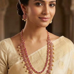 May include: A two-layered gold necklace featuring floral-shaped motifs set with oval-cut pink stones and small dangling cream-colored pearls. Matching floral-shaped drop earrings with pink stones and cream-colored pearls complete the jewelry set.