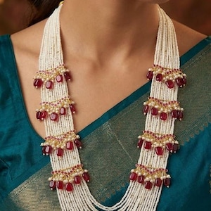 May include: A multi-strand necklace featuring numerous rows of small white seed beads. The necklace is accented with four symmetrical tiers of deep red, oval-shaped stones and small white pearls, arranged in horizontal segments along the strands.