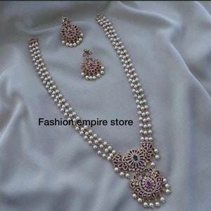 May include: A three-strand pearl necklace with a gold pendant featuring a floral design with pink, green, and white stones. Matching earrings with a similar design are also included. The necklace is on a white fabric background.
