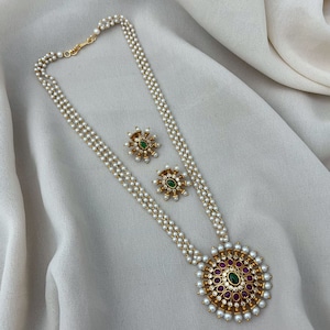 Puede incluir: Un collar de perlas blancas con un colgante de oro que presenta una piedra preciosa verde y rosa rodeada de pequeñas perlas blancas. El collar está formado por tres hileras de perlas y viene con pendientes a juego.