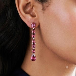 Pode incluir: Um brinco longo pendente com cinco pedras preciosas cor-de-rosa facetadas cravadas em metal prateado. O design consiste em pedras ovais e em forma de gota dispostas verticalmente, terminando numa pedra maior na parte inferior da peça.