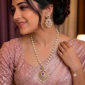 May include: A gold-toned necklace and matching drop earrings set featuring white pearls and small clear stones arranged in a floral pattern, with a central teardrop-shaped pink stone in each piece, worn against a shimmering light pink fabric.