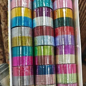 May include: A collection of thin metal bangles arranged in three vertical columns within a white rectangular box. The bangles feature a variety of metallic and glossy colors including red, blue, yellow, green, purple, silver, pink, and gold.