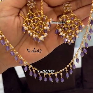 May include: A gold-toned jewelry set featuring a necklace and matching earrings. The pieces are adorned with small white pearls and purple teardrop-shaped crystals. The earrings have a scalloped lattice design. Text reads Beautiful, *s d143, *s 00997.