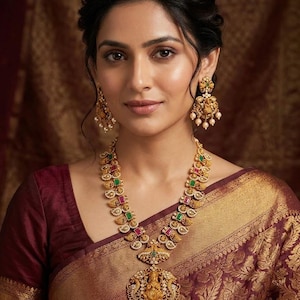 May include: A gold-toned necklace and matching drop earrings set featuring intricate metalwork, embedded with rectangular green and pink stones, and accented with small white pearls. The central pendant displays a detailed deity figure.
