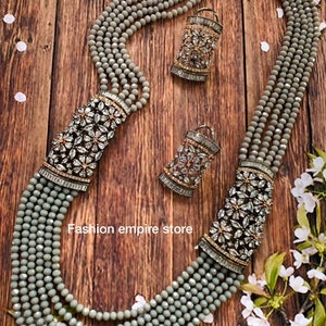 Pode incluir: Um colar de várias voltas feito de contas cinzas com dois brincos pendentes combinando. Os pingentes são de prata com strass transparentes em um design floral. O colar e os brincos estão sobre uma superfície de madeira com flores brancas.