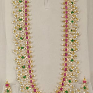 May include: A gold-toned necklace and earring set. The necklace features a central row of rectangular pink stones, interspersed with green and white accents. The earrings mirror the necklace design. Small white pearls and gold details complete the set.