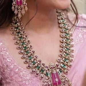 May include: A gold-toned necklace and matching chandelier earrings set featuring large rectangular pink stones, smaller oval green stones, and clear pavé-set crystals. The jewelry displays an intricate metalwork design with dangling elements.