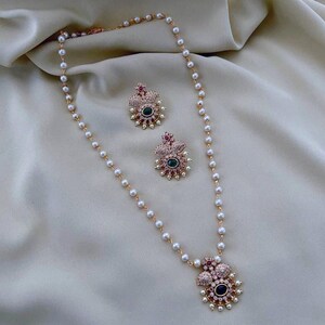 May include: A gold necklace with a pearl chain and a floral pendant featuring green and red stones. Matching earrings with the same design are also included.
