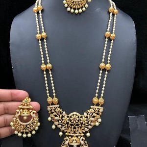 Puede incluir: Un collar de oro con una cadena de perlas y un gran colgante con intrincados diseños florales y piedras blancas. El colgante está unido a un colgante más pequeño con forma de media luna. El collar viene con pendientes a juego.