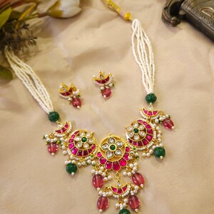May include: A gold-toned necklace and earring set featuring a floral design with pink and green gemstones. The necklace has a multi-strand white beaded chain. The earrings mirror the necklace's design. The jewelry set is displayed on a neutral background.