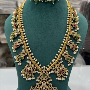 May include: Gold-coloured necklace and matching earrings set. The necklace features intricate filigree designs, adorned with red and green gemstones, and small white pearls. The pendant has a large, central red stone. The earrings mirror the necklace's design.