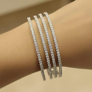 May include: Four silver bracelets, each encrusted with small, sparkling gemstones, are displayed. The bracelets are arranged side by side, showcasing their intricate design and the brilliance of the stones. The bracelets are on a light skin tone.