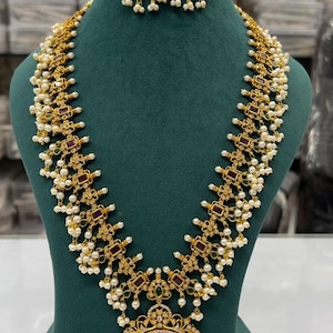 以下が含まれることがあります： 赤い石が小さな石で囲まれた大きなペンダントが付いた金のネックレス。ネックレスは白い真珠と緑の石で飾られています。赤い石と緑の石がついたマッチングイヤリングも含まれています。