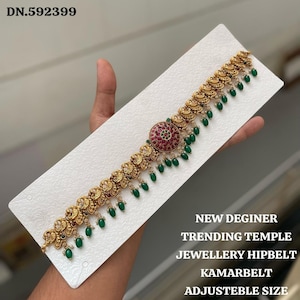 Könnte beinhalten: Goldfarbener Hüftgürtel mit einem zentralen runden Design aus roten und grünen Edelsteinen. Der Gürtel hat aufwendige Details und hängende grüne Perlen. Der Text auf dem Bild lautet "NEW DEGINER TRENDING TEMPLE JEWELLERY HIPBELT KAMARBELT ADJUSTEBLE SIZE."
