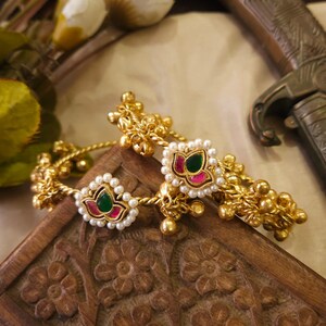 May include: Gold-toned bangles with intricate detailing. Each bangle features a central floral design with a green and red center, surrounded by small white pearls. The bangles rest on a carved wooden surface.