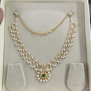 以下が含まれることがあります： 緑色の石のペンダントと白い真珠のアクセントが付いた金のネックレス。ネックレスは、白い箱に白いベルベットのクッション2つと一緒に展示されています。