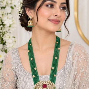 May include: A long necklace featuring multiple strands of small green beads accented with small gold floral spacers and a large central pendant with a rectangular red stone. Matching bell-shaped jhumka earrings with green beads.