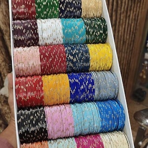 May include: A box filled with multiple stacks of colorful bangles. The bangles are in various colors, including red, green, blue, gold, and pink. Some bangles have a glittery design. The box is white and angled.
