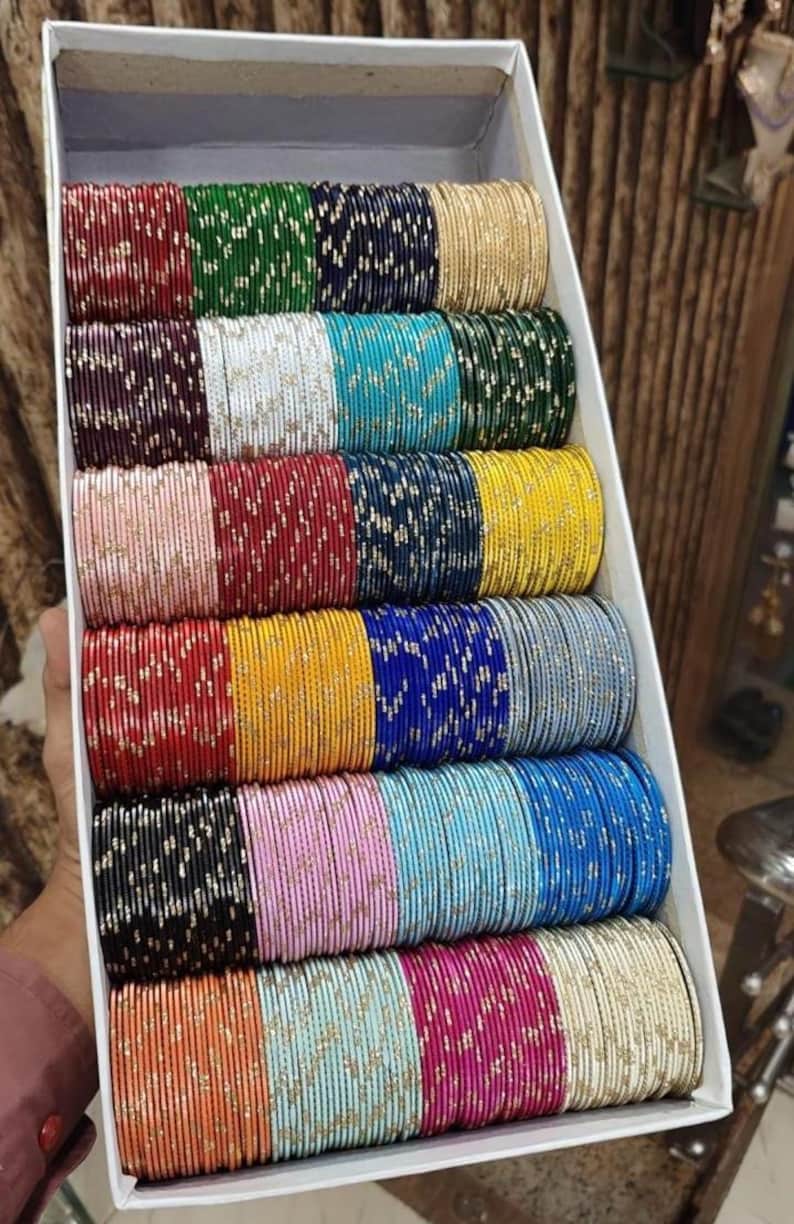 May include: A box filled with multiple stacks of colorful bangles. The bangles are in various colors, including red, green, blue, gold, and pink. Some bangles have a glittery design. The box is white and angled.