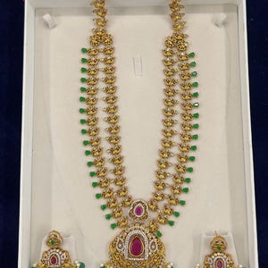 Op de afbeelding: Een gouden ketting met een hanger met een roze steen en groene accenten. De ketting wordt gepresenteerd in een witte doos met bijpassende oorbellen.
