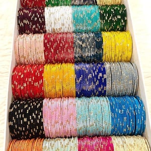 May include: A box filled with stacks of colorful bangles. The bangles come in a variety of colors, including red, green, blue, yellow, pink, and gold. Each bangle is adorned with a delicate pattern.