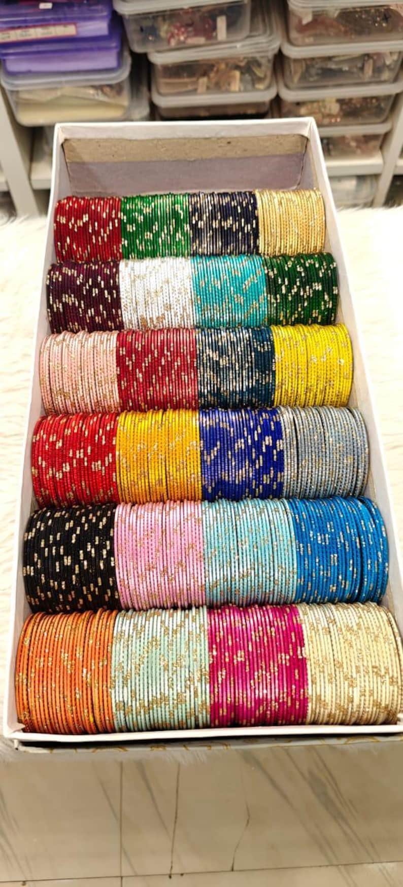 May include: A box filled with stacks of colorful bangles. The bangles come in a variety of colors, including red, green, blue, yellow, pink, and gold. Each bangle is adorned with a delicate pattern.