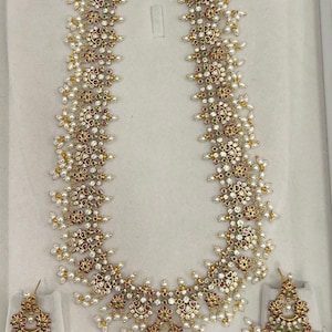 May include: A long gold necklace featuring intricate floral-inspired metalwork set with small white stones, red and green accents, and dangling white pearls. The set includes a pair of matching triangular earrings with similar stone and pearl details.