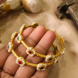 May include: Two gold bangles with floral designs. Each flower has a red or green center surrounded by small white pearls. The bangles are held in a hand, showcasing the intricate details and craftsmanship of the jewelry.