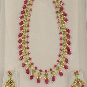 May include: A gold necklace and earrings set with pink and green stones. The necklace has a long chain with multiple strands of pink oval stones and green leaves. The earrings are dangle style with pink oval stones and green leaves.