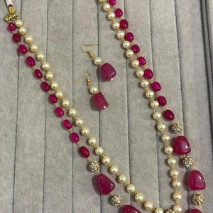 May include: A two-strand necklace and matching drop earrings set featuring alternating cream-colored pearls and smooth, deep pink oval beads. The jewelry includes small gold-toned metal accents between the beads and gold-toned earring hooks.