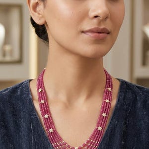May include: A multi-strand necklace featuring five rows of small, faceted, deep pink ruby-colored beads. The strands are accented with small, gold-toned, star-shaped spacers and a single, centered, cream-colored pearl on each of the five strands.