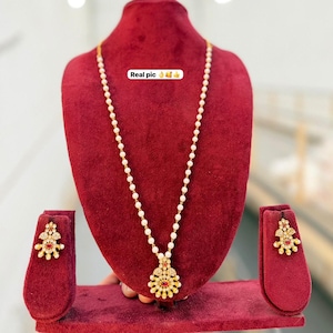 May include: A gold-toned necklace featuring a single strand of white pearls with a central pendant containing a red stone and clear accents, accompanied by matching stud earrings. The text "Real pic" is visible on the necklace display.