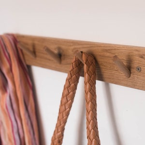 May include: A wooden wall-mounted coat rack with four hooks. The rack is made of light brown wood and has a natural finish. A striped scarf and two leather straps are hanging on the hooks.