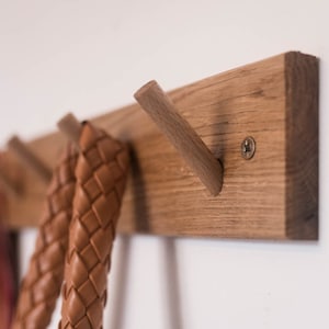 May include: A wooden wall-mounted coat rack with four hooks. The rack is made of light brown wood and has a natural finish. Two brown leather straps are hanging from the hooks.