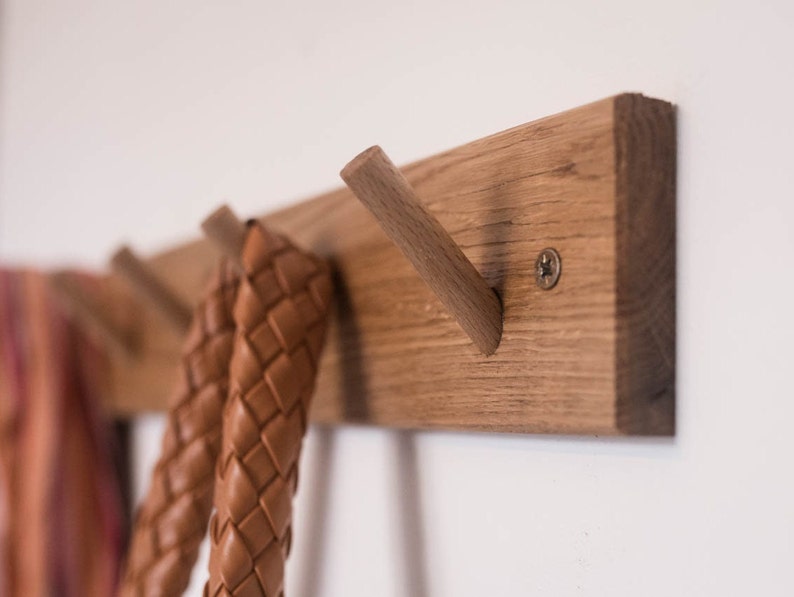 May include: A wooden wall-mounted coat rack with four hooks. The rack is made of light brown wood and has a natural finish. Two brown leather straps are hanging from the hooks.