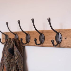 May include: A wooden coat rack featuring five antique-style metal hooks. The hooks are attached to a light brown wooden board. A patterned scarf hangs from some of the hooks. The rack is mounted on a white wall.