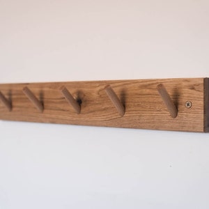 May include: A wooden wall-mounted coat rack with five hooks. The rack is made of natural wood with a light brown finish. The hooks are made of the same wood and are shaped like a V.