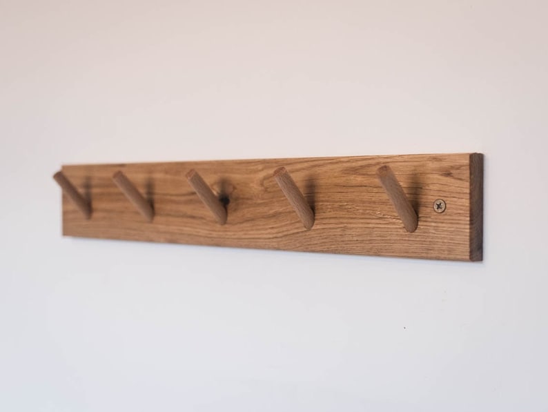 May include: A wooden wall-mounted coat rack with five hooks. The rack is made of natural wood with a light brown finish. The hooks are made of the same wood and are shaped like a V.
