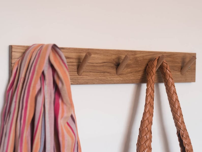 May include: A wooden wall-mounted coat rack with four hooks. A striped scarf hangs on one hook and a brown leather bag hangs on another hook.