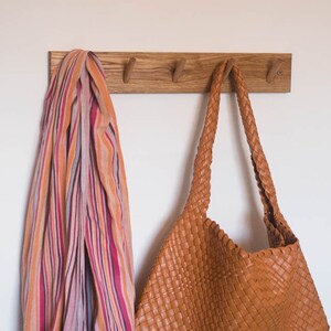 May include: A wooden wall-mounted coat rack with four hooks. A striped scarf and a brown leather bag are hanging on the hooks.
