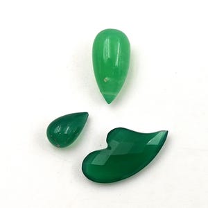 May include: Three polished green gemstones of varying shapes and sizes. One is a smooth, light green teardrop cabochon, another is a smaller, darker green teardrop, and the third is a larger, faceted, dark green freeform stone with a curved edge.