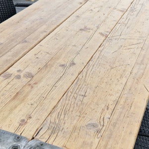 Op de afbeelding: Een close-up van een houten tafelblad met een natuurlijke houtkleur. Het tafelblad is gemaakt van verschillende planken hout die met metalen beugels aan elkaar zijn bevestigd. De beugels hebben een zilveren afwerking en zijn vastgeschroefd. Het tafelblad is licht verweerd en heeft een rustieke uitstraling.