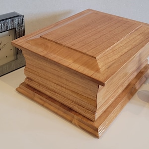 May include: A wooden box with a hinged lid and a decorative design. The box is made of light brown wood with a natural grain pattern. The box is closed and sitting on a white surface.