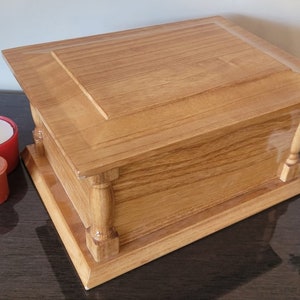 May include: A wooden box with a hinged lid and decorative trim. The box is made of light brown wood and has a natural wood finish. Three red candles are in the foreground.