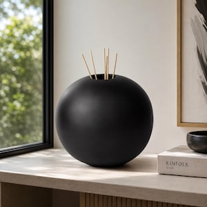 May include: A spherical, matte black vase with several light brown reeds. The vase is on a light-coloured surface near a window. The vase is a decorative home accent, suitable for a living room or bedroom.