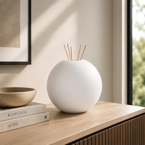 May include: A white, spherical diffuser with several brown reeds. The diffuser is on a wooden surface next to a bowl and two books. The books are titled "KINFOLK" and "at 100".