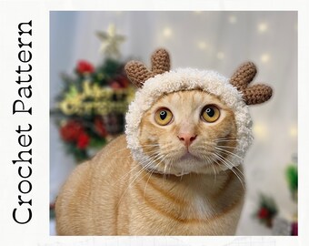 Puede incluir: Primer plano de un gato jengibre con un gorro de reno de ganchillo blanco y marrón. El gato mira directamente a la cámara con una expresión curiosa.
