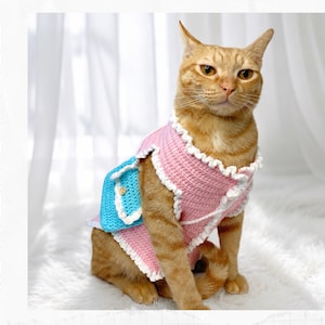 May include: A ginger cat wearing a pink crochet vest with white ruffled trim and a small blue crochet pocket featuring a wooden button. The text Crochet Pattern is printed vertically on the left side of the image in black capital letters.